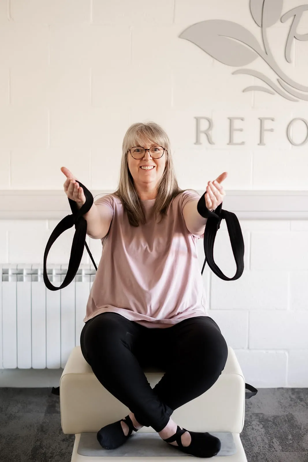 Smiling client holding Reformer straps with open arms under the Pilates Flow Reformer logo